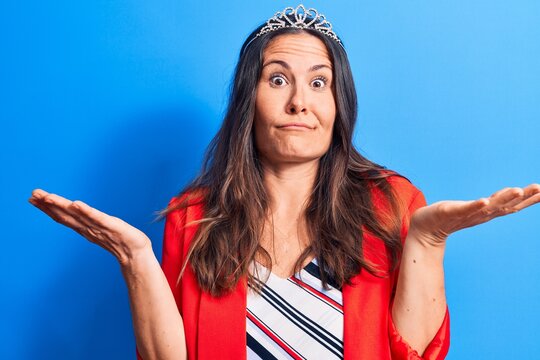 Young beautiful brunette woman wearing princess crown standing over blue background clueless and confused with open arms, no idea and doubtful face.