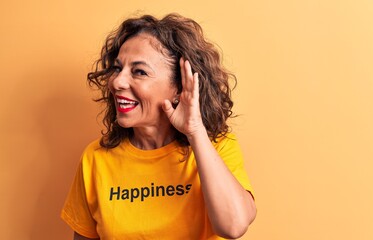 Middle age beautiful woman wearing t-shirt with happiness word over yellow background smiling with hand over ear listening and hearing to rumor or gossip. Deafness concept.