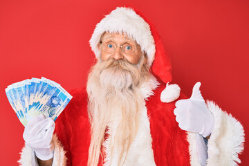 Old senior man with grey hair and long beard wearing santa claus costume holding african rands smiling happy and positive, thumb up doing excellent and approval sign