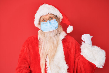 Old senior man wearing santa claus costume wearing safety mask pointing thumb up to the side smiling happy with open mouth