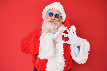 Old senior man wearing santa claus costume and sunglasses smiling in love showing heart symbol and shape with hands. romantic concept.