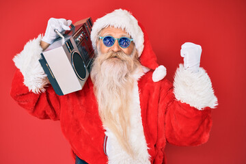 Old senior man wearing santa claus costume and boombox screaming proud, celebrating victory and success very excited with raised arms