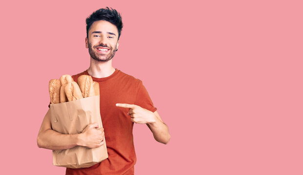 Young hispanic man holding paper bag with bread smiling happy pointing with hand and finger