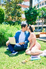 Beautiful couple kissing sitting on the grass using laptop at park