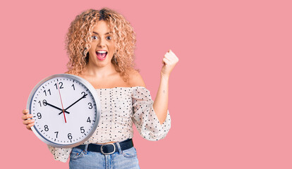 Young blonde woman with curly hair holding big clock screaming proud, celebrating victory and success very excited with raised arms