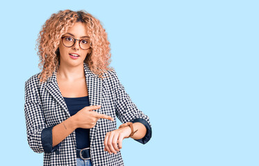 Young blonde woman with curly hair wearing business jacket and glasses in hurry pointing to watch time, impatience, upset and angry for deadline delay