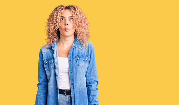 Young blonde woman with curly hair wearing casual denim jacket making fish face with lips, crazy and comical gesture. funny expression.