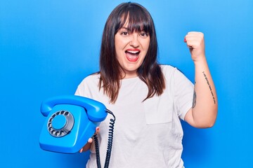 Young plus size woman holding vintage telephone screaming proud, celebrating victory and success very excited with raised arms