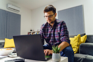 Young caucasian man working on the laptop computer at home - Male accountant sitting on the couch in his apartment work late at night alone - business and finance concept