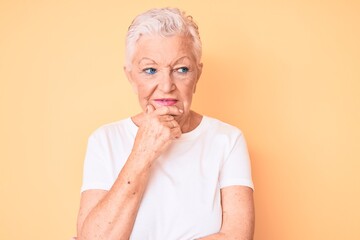 Senior beautiful woman with blue eyes and grey hair wearing classic white tshirt over yellow background with hand on chin thinking about question, pensive expression. smiling with thoughtful face.