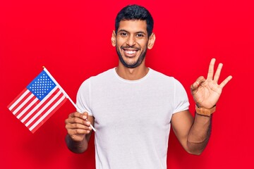 Young latin man holding united states flag doing ok sign with fingers, smiling friendly gesturing...