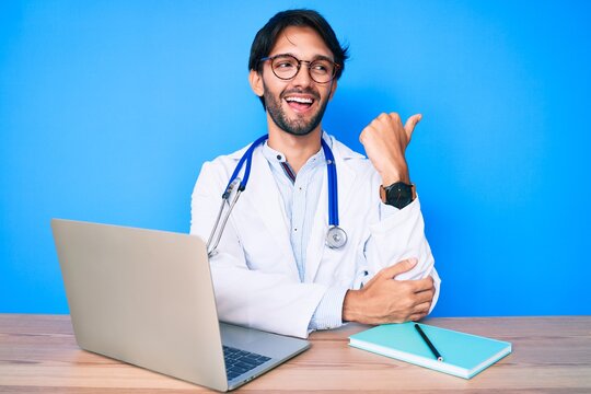 Handsome hispanic man wearing doctor uniform working at the clinic pointing thumb up to the side smiling happy with open mouth