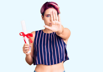 Young beautiful woman with pink hair holding graduate degree diploma with open hand doing stop sign with serious and confident expression, defense gesture