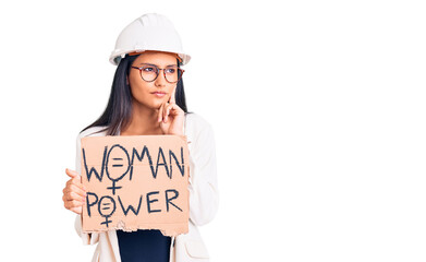 Young beautiful latin girl wearing architect hardhat holding woman power banner serious face thinking about question with hand on chin, thoughtful about confusing idea
