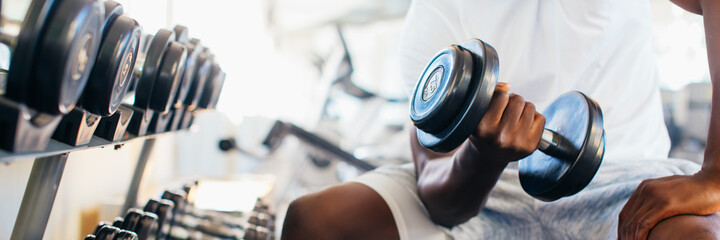 Young African American man sitting and lifting a dumbbell close to the rack at gym. Male weight training person doing a biceps curl in fitness center