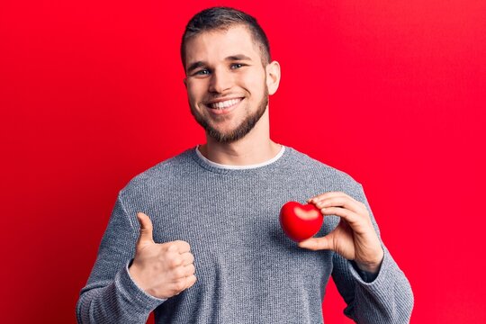 Young handsome man holding heart smiling happy and positive, thumb up doing excellent and approval sign