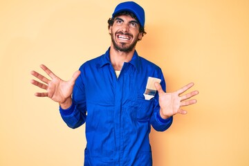 Handsome young man with curly hair and bear wearing builder jumpsuit uniform crazy and mad shouting and yelling with aggressive expression and arms raised. frustration concept.