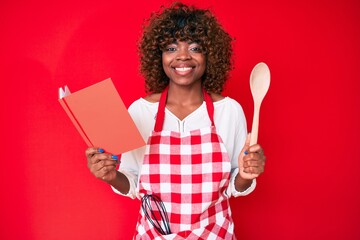 Young african american woman wearing professional baker apron reading cooking recipe book looking positive and happy standing and smiling with a confident smile showing teeth