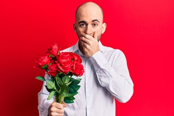 Young handsome bald man holding bouquet of red flowers covering mouth with hand, shocked and afraid...