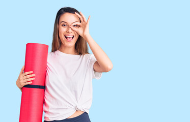Young beautiful girl holding yoga mat smiling happy doing ok sign with hand on eye looking through...