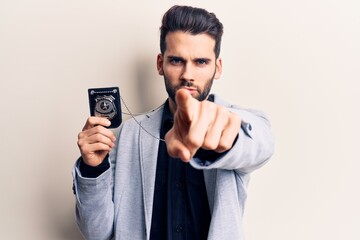 Young handsome man with beard holding police badge pointing with finger to the camera and to you, confident gesture looking serious