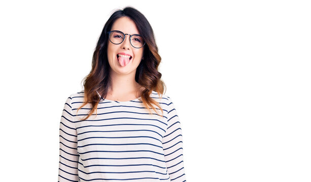 Beautiful young brunette woman wearing casual clothes and glasses sticking tongue out happy with funny expression. emotion concept.