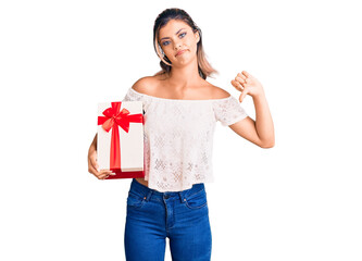 Young beautiful woman holding gift with angry face, negative sign showing dislike with thumbs down, rejection concept