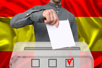 male voter drops a ballot in a transparent ballot box against the background of the national flag of Spain, concept of state elections, referendum