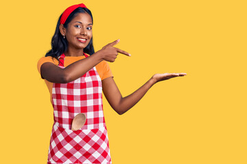Fototapeta na wymiar Young indian girl wearing professional baker apron amazed and smiling to the camera while presenting with hand and pointing with finger.