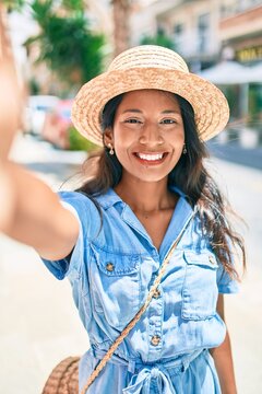 Young beautiful indian woman wearing summer hat smiling happy making selfie by the camera at the city.