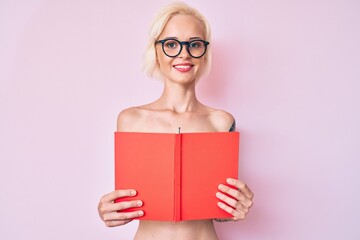 Young blonde woman with tattoo standing shirtless reading book smiling with a happy and cool smile on face. showing teeth.