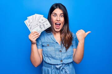 Young beautiful brunette woman holding dollars banknotes over isolated blue background pointing thumb up to the side smiling happy with open mouth