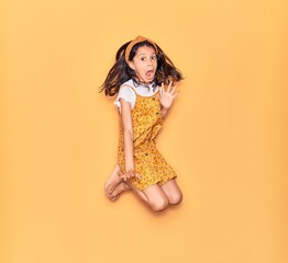 Adorable hispanic child girl scared wearing casual dress and diadem. Jumping over isolated yellow...