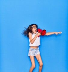 Adorable hispanic child girl wearing glasses. Screaming using megaphone jumping over isolated blue background