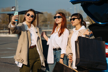 Young women at the car with shopping bags. Girls take selfie