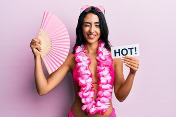 Beautiful hispanic woman wearing bikini and handfan holding hot word winking looking at the camera...