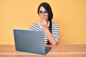 Brunette teenager girl working using computer laptop covering mouth with hand, shocked and afraid for mistake. surprised expression