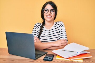 Brunette teenager girl working at the office with laptop happy face smiling with crossed arms looking at the camera. positive person.