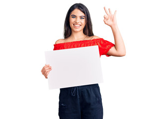 Brunette teenager girl holding blank empty banner doing ok sign with fingers, smiling friendly gesturing excellent symbol
