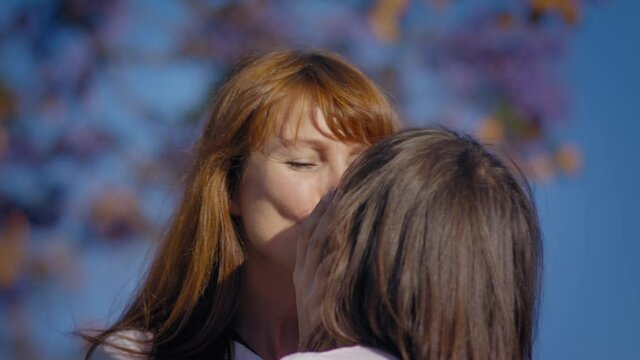 Adult kissing a child.
Mom hugs daughter.
Jacaranda and blue sky on background.
White woman touching girl's hair.
Female smile. Shot URSA 4.6K
