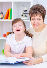 A girl with down syndrome with thick dark hair is sitting next to her teacher holding a book in her hands and smiling while looking at the camera. Education accessible to children with disabilities co