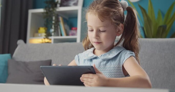Portrait of pleasant young girl in stylish dress using digital tablet for drawing. Beautiful kid making paintings on modern gadget while staying at home.