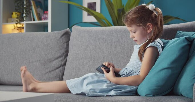 Side view of small kid with two ponytails lying on grey couch and playing on modern tablet. Pretty girl with barefoot relaxing at home and using digital gadget.