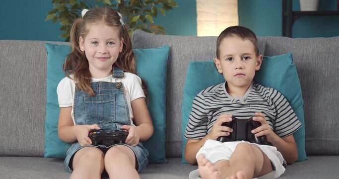 Joyful girl and boy in casual clothing sitting on grey couch and using joysticks for playing video games. Sister and brother relaxing together at home and taking fun.