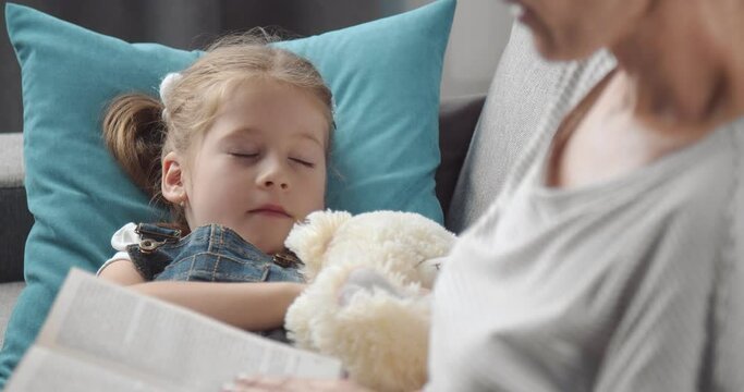 Pretty girl lying on couch with teddy bear and fell asleep while her mother reading book. Woman taking care of her little child while staying at home.