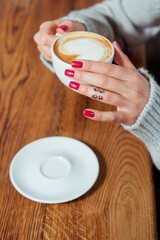 Young woman holding cup of coffee with latte art. She is wearing gray wool sweater and trendy silver ring. Morning in a cafe.