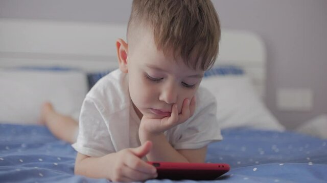 Cute little boy preschool child using a phone or digital tablet technology device looks telephone,plays in the phone sitting rest lying on sofa alone.Children tech addiction concept play game at home.