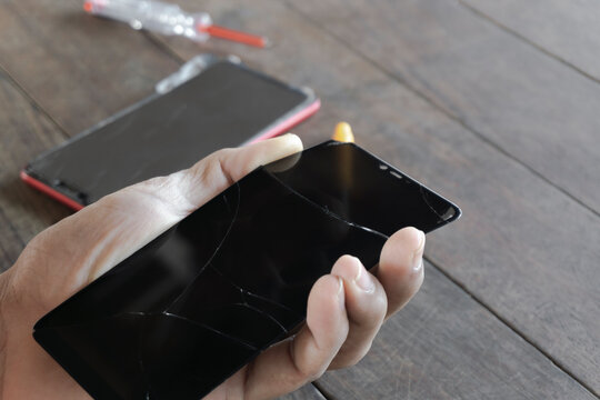 A Man Technician Holds A Black Smartphone Screen Cracked Or Broken From A Drop For Repair Or Cell Phone Replacement, With A Red Phone Body On A Wooden Table With A Screwdriver.