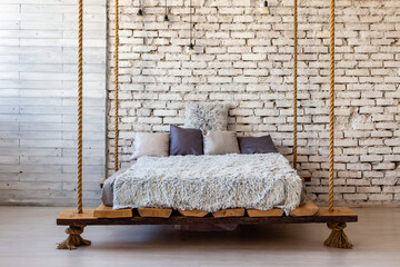 Wooden bed with pillows and a fur blanket hanging on the ropes in the loft interior of a stylish modern bedroom.