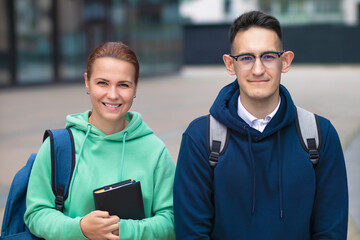 Portrait of couple of two beautiful happy university or college students, guy in glasses and young woman with book or textbook. Smiling pupils outdoors at campus with backpacks. Boy and girl